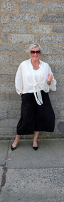 A woman stands against a gray stone wall wearing a white long sleeve tie front blouse, black wide leg cropped pants, and black pointed flats.