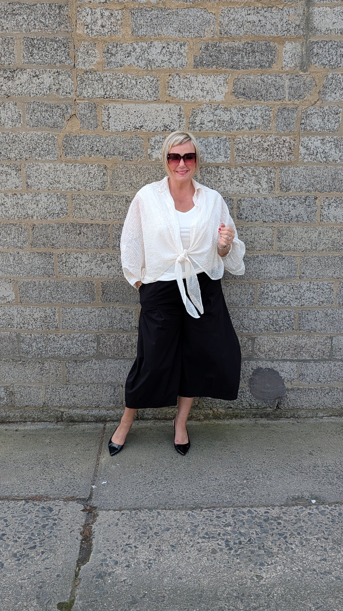 A woman stands against a gray stone wall wearing a white long sleeve tie front blouse, black wide leg cropped pants, and black pointed flats.