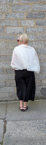 A woman stands with her back to the camera against a grey stone wall, wearing a white textured wrap or shawl draped over her shoulders, a black wide leg cropped skirt, and black strappy heels.