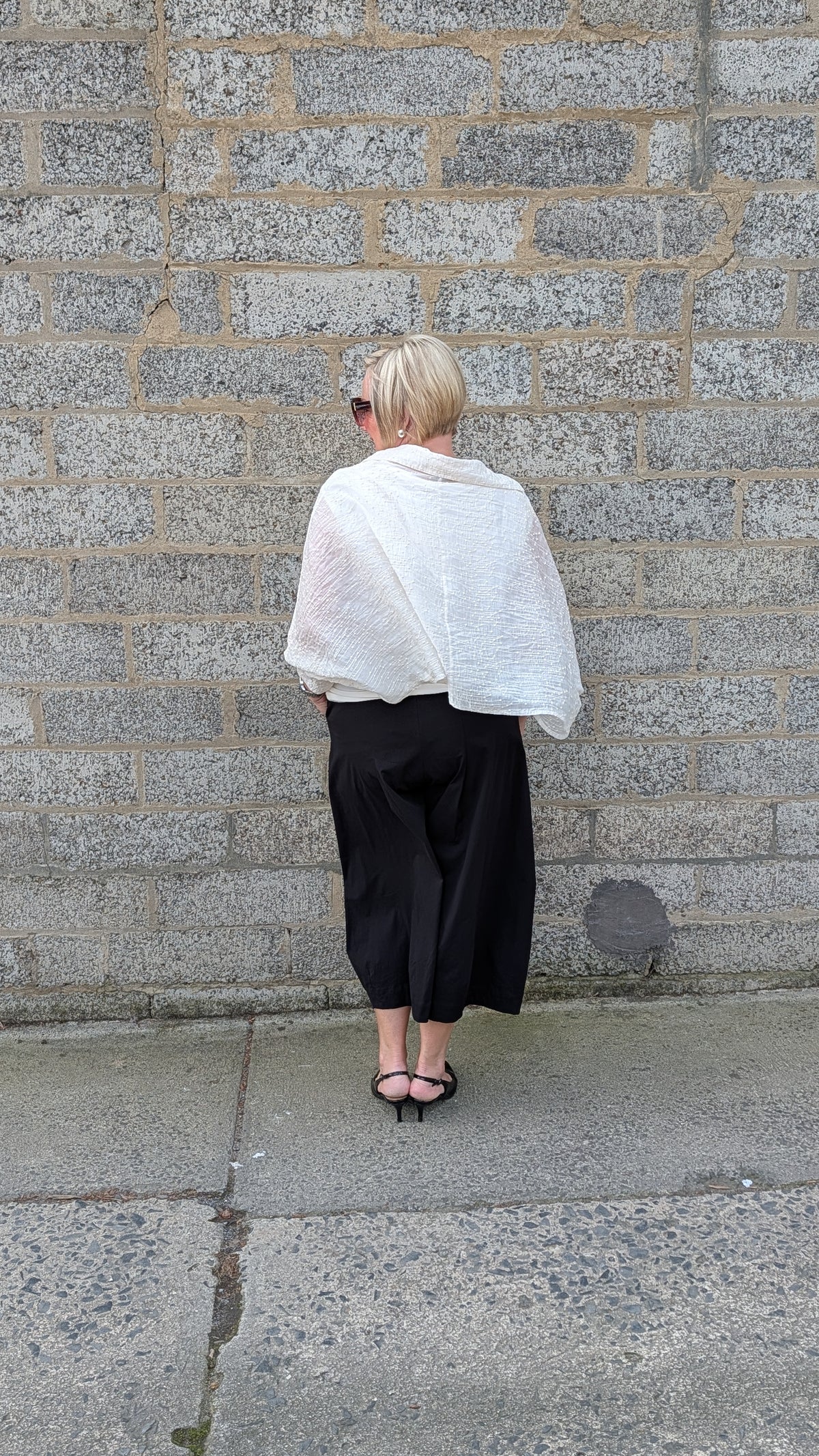 A woman stands with her back to the camera against a grey stone wall, wearing a white textured wrap or shawl draped over her shoulders, a black wide leg cropped skirt, and black strappy heels.