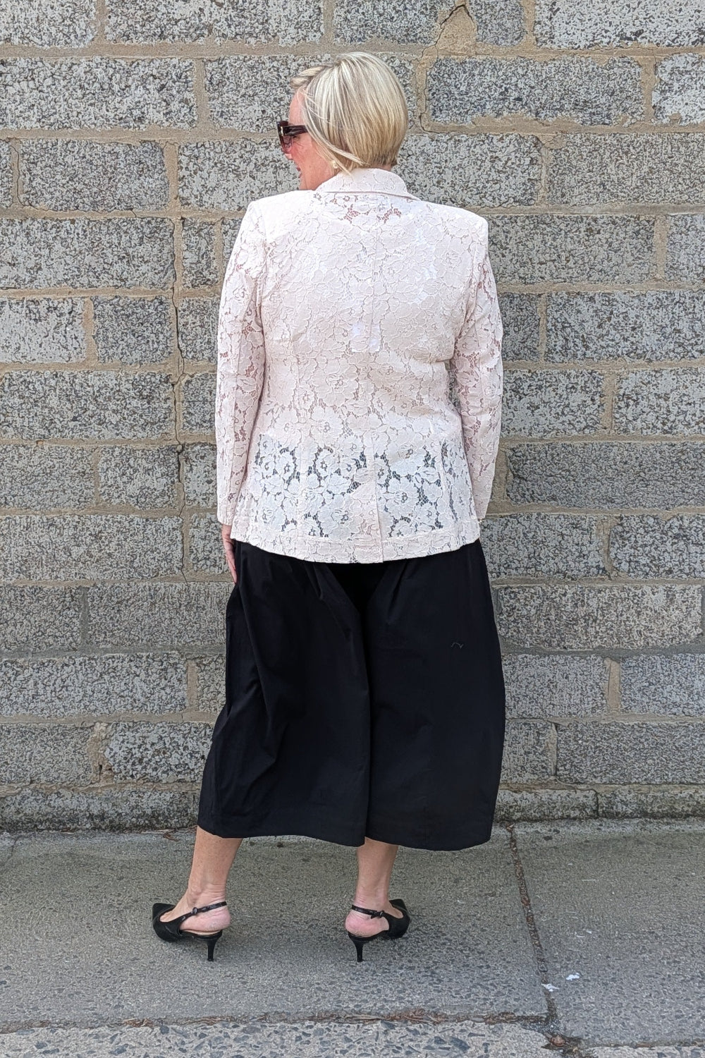 Lace Blazer by Two-Ts in Straw back view lstraw lace  jacket and black wide-leg cropped pants against a gray stone wall.