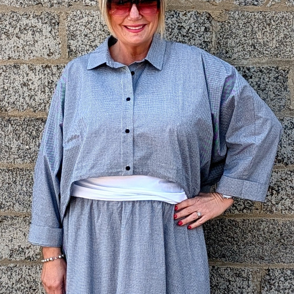 A woman wearing a light blue-gray striped maxi shirt dress with a white undershirt and white pointed-toe heels, standing against a gray stone wall.