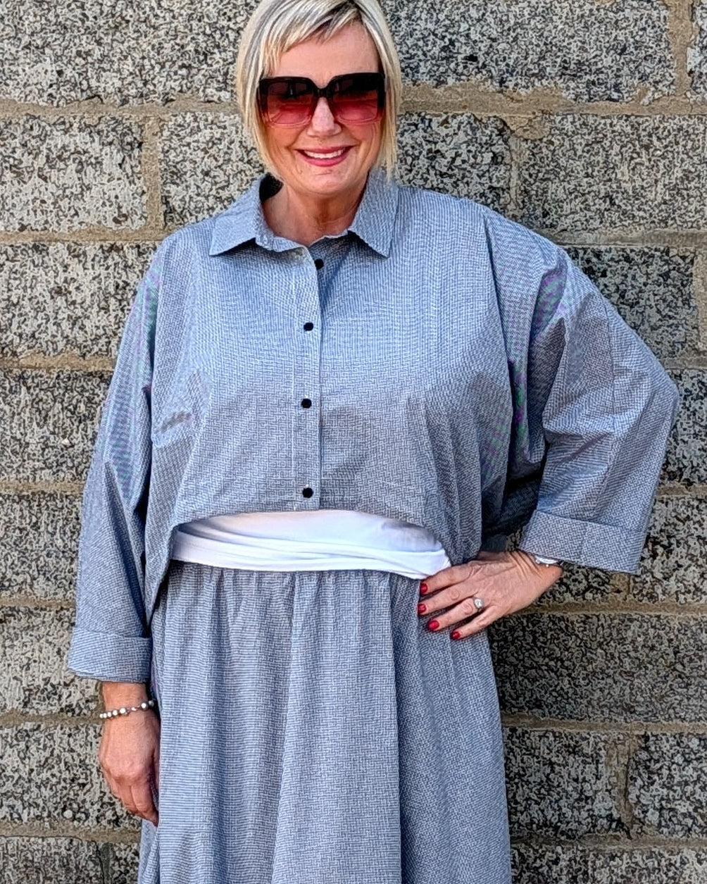 A woman wearing a light blue-gray striped maxi shirt dress with a white undershirt and white pointed-toe heels, standing against a gray stone wall.