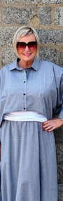 A woman wearing a light blue-gray striped maxi shirt dress with a white undershirt and white pointed-toe heels, standing against a gray stone wall.