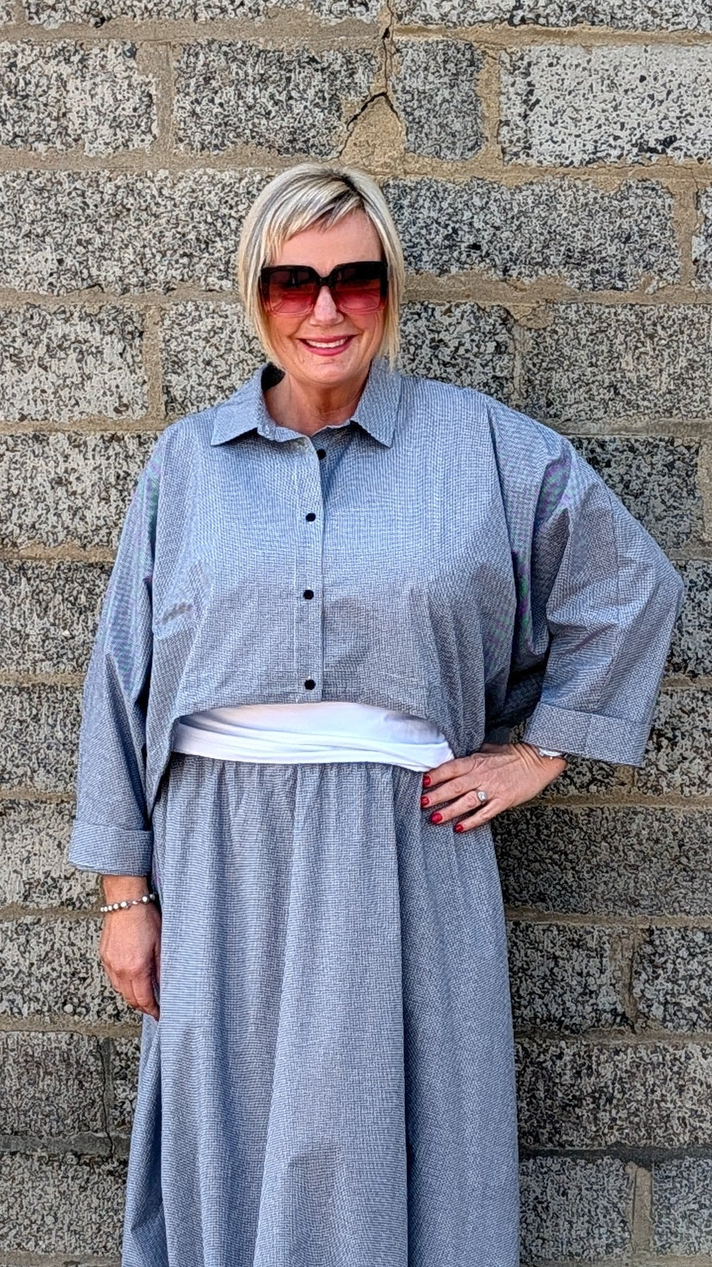 A woman wearing a light blue-gray striped maxi shirt dress with a white undershirt and white pointed-toe heels, standing against a gray stone wall.