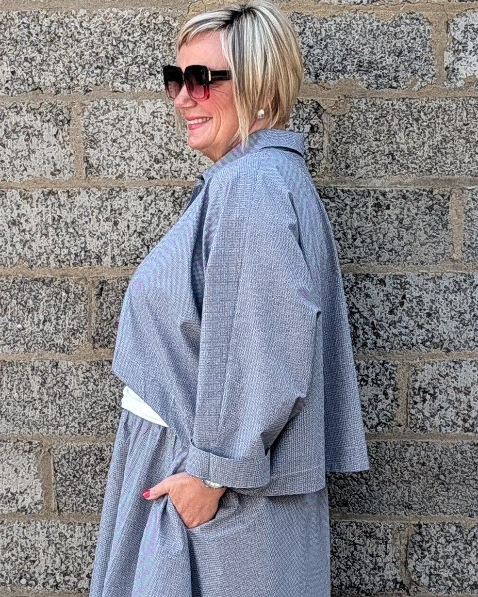 A woman with short blonde hair and sunglasses stands in profile against a gray stone wall, wearing a loose, oversized light gray poncho-style garment with white shoes.