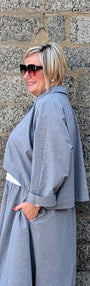 A woman with short blonde hair and sunglasses stands in profile against a gray stone wall, wearing a loose, oversized light gray poncho-style garment with white shoes.
