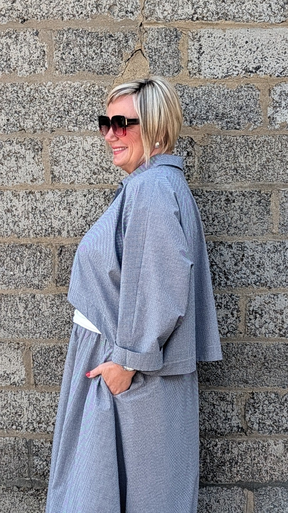 A woman with short blonde hair and sunglasses stands in profile against a gray stone wall, wearing a loose, oversized light gray poncho-style garment with white shoes.