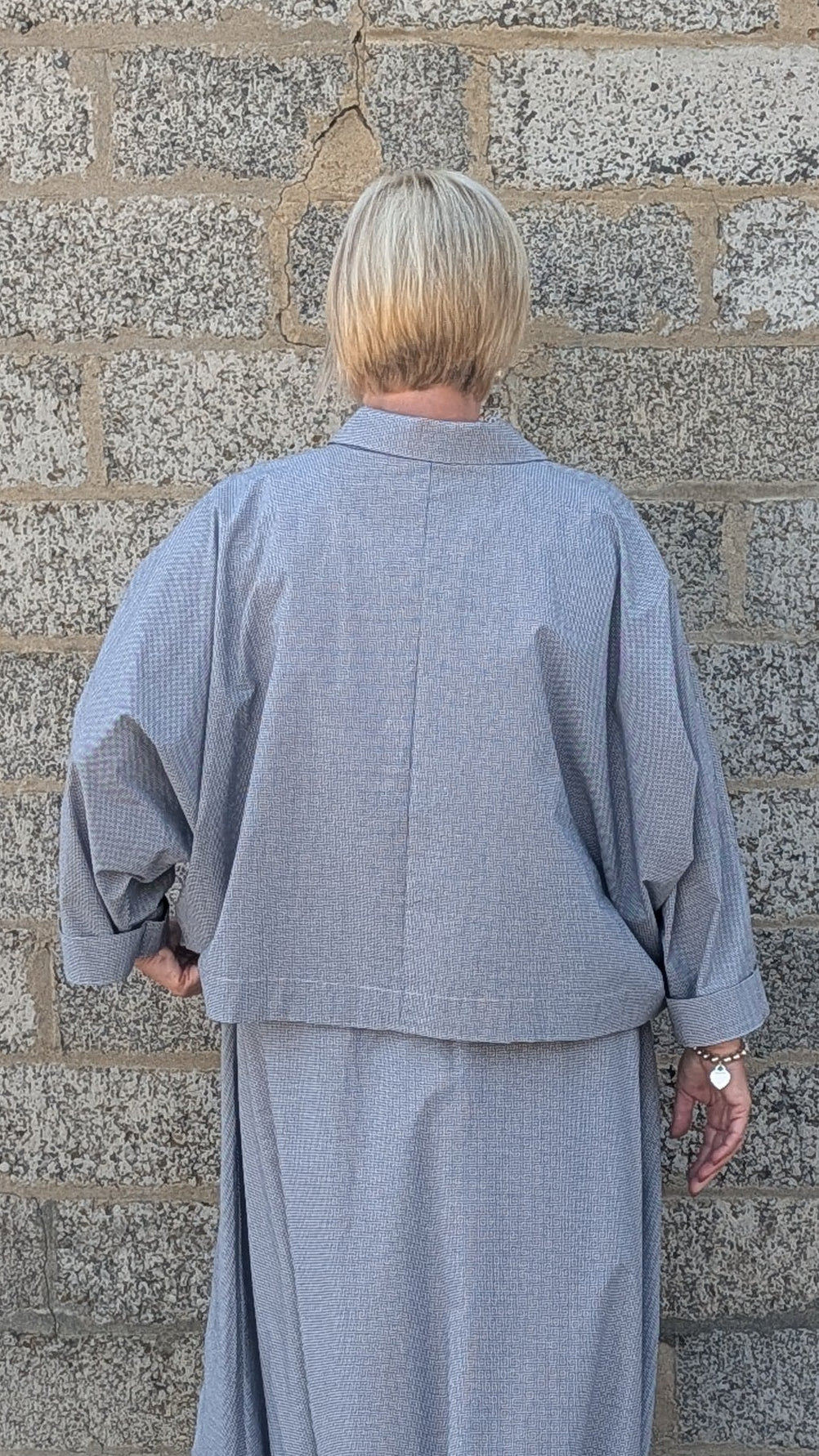 A person viewed from behind wearing a light gray two-piece outfit consisting of a cropped long-sleeve top and a matching maxi skirt, standing against a gray stone wall.