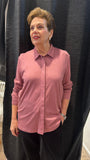An mature woman with short brown hair stands in a fitting room wearing a dusty rose pink long-sleeve button-up shirt and black pants, posing in front of a black curtain.