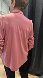 A woman viewed from behind wearing a dusty rose pink long-sleeve collared shirt with ruched sleeves, paired with black pants in a fitting room or boutique setting.