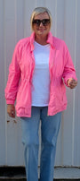 A woman wearing a bright pink lightweight zip-up jacket over a white t-shirt, light blue jeans, and white sneakers, standing in front of a corrugated metal wall.