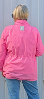 A person viewed from behind wearing a bright pink lightweight jacket, light blue jeans, and white sneakers, standing in front of a corrugated white metal wall.
