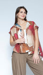 A woman modeling a short sleeve button up shirt with an abstract multicolor print in shades of red, beige, brown, and blue, worn over a white top and paired with brown pants, against a light gray studio background.