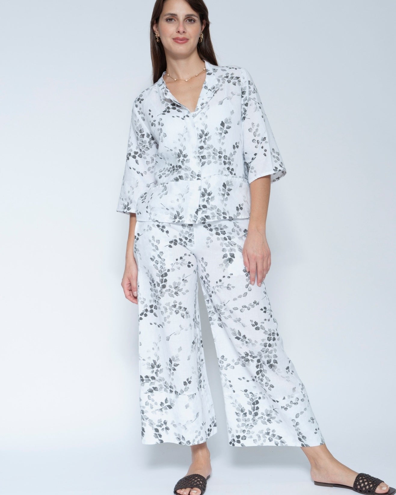  Front view  matching two piece jacket and pant set with a white base and black floral print, featuring a short sleeve button up top and wide leg pants, paired with black slide sandals.