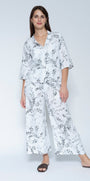  Front view  matching two piece jacket and pant set with a white base and black floral print, featuring a short sleeve button up top and wide leg pants, paired with black slide sandals.